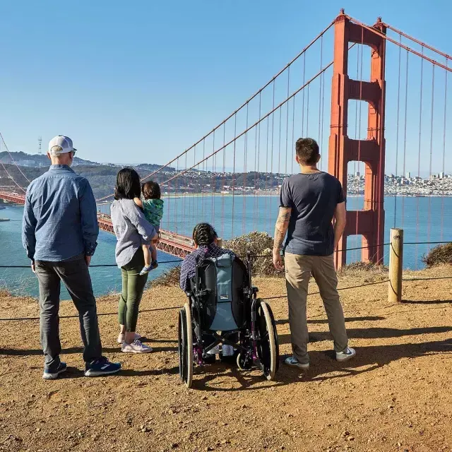 A group of people, including one person in a wheelchair, is seen from behind as they look at the Golden Gate Bridge from the Marin Headlands.